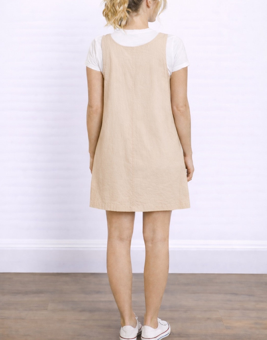 Back view of woman wearing a beige Sleeveless Suspender Shift Dress over a white shirt on a plain background.