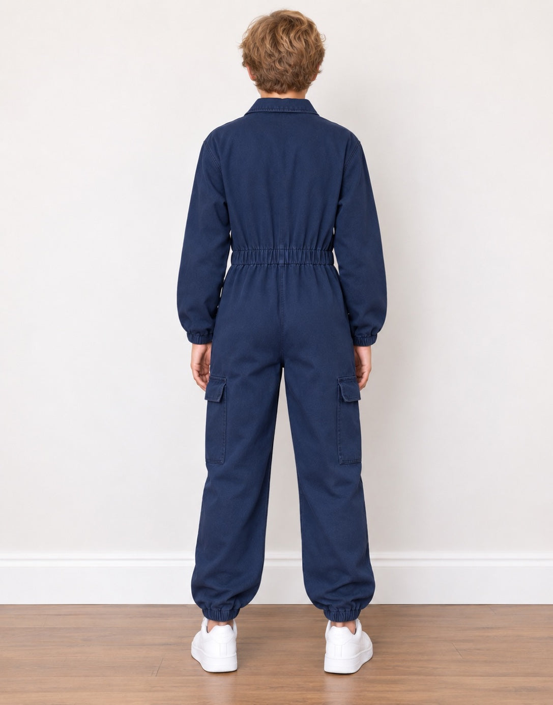 A young boy from the back wearing a dark blue denim jumpsuit with a gathered elastic waist, long sleeves, and cargo pockets, paired with white sneakers against a plain off-white wall.