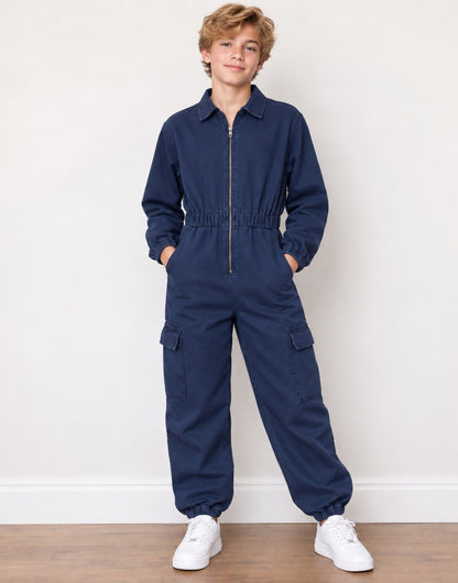 A young boy stands wearing a dark blue denim jumpsuit with a front zipper, elastic waist, and cargo pockets, paired with white sneakers against a plain white wall and wood floor.