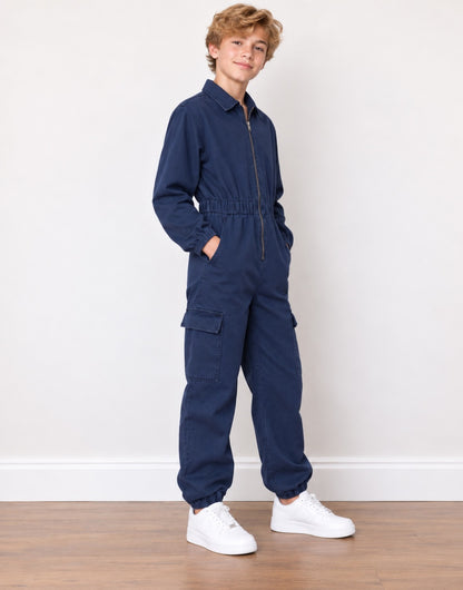 Side view of a young boy stands wearing a dark blue denim jumpsuit with white sneakers against a plain off-white wall and wooden floor.