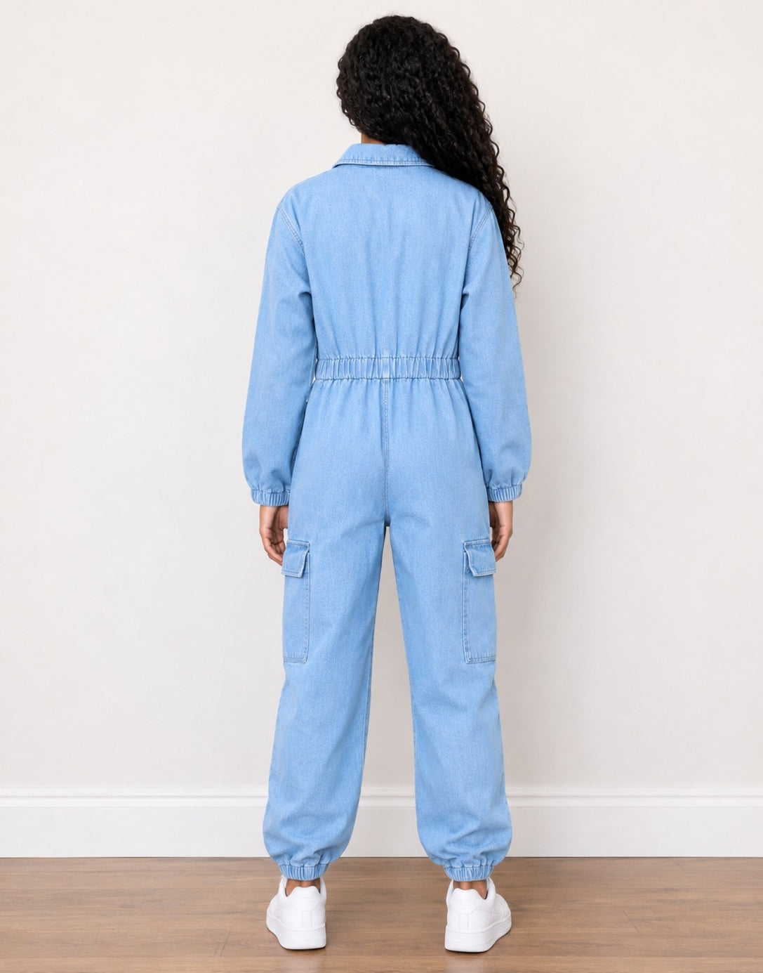 A young girl from the back wearing a light blue denim jumpsuit with a gathered elastic waist, long sleeves, and cargo pockets, paired with white sneakers against a plain off-white wall.