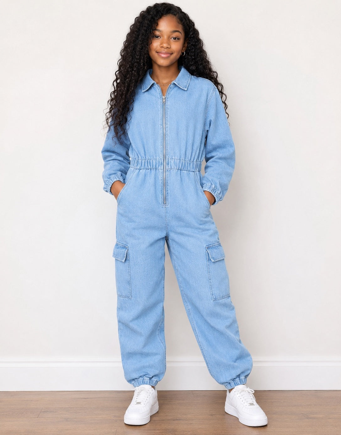 A young girl with long curly black hair stands wearing a light blue denim jumpsuit with a front zipper, elastic waist, and cargo pockets, paired with white sneakers against a plain white wall and wood floor.