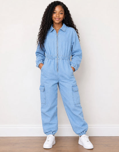 A young girl with long curly black hair stands wearing a light blue denim jumpsuit with a front zipper, elastic waist, and cargo pockets, paired with white sneakers against a plain white wall and wood floor.