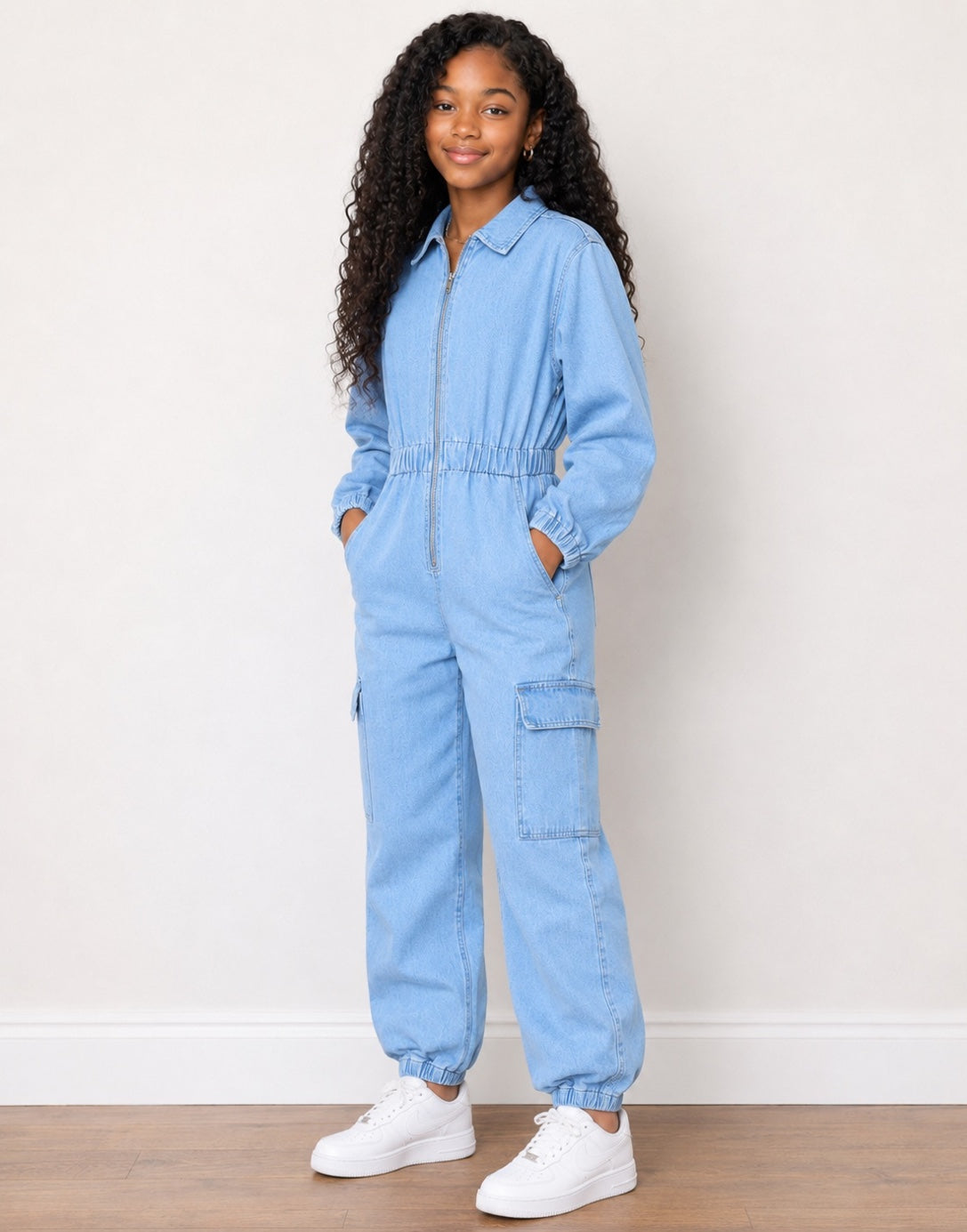 Side view of a young girl with long curly black hair stands wearing a light blue denim jumpsuit with white sneakers against a plain off-white wall and wooden floor.