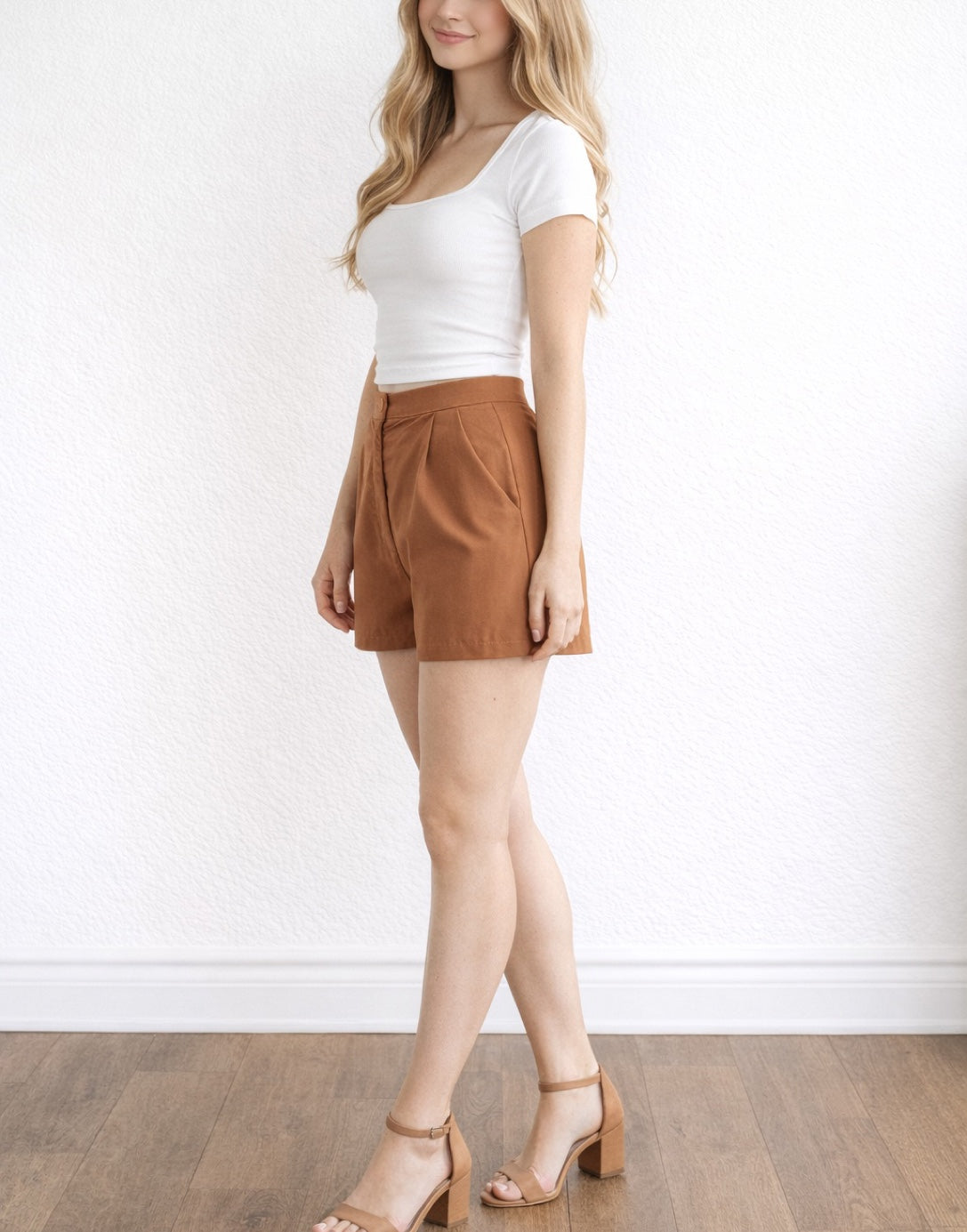 Side view of woman wearing a white top and brown Wide Leg Pleats Shorts standing in a plain white background.