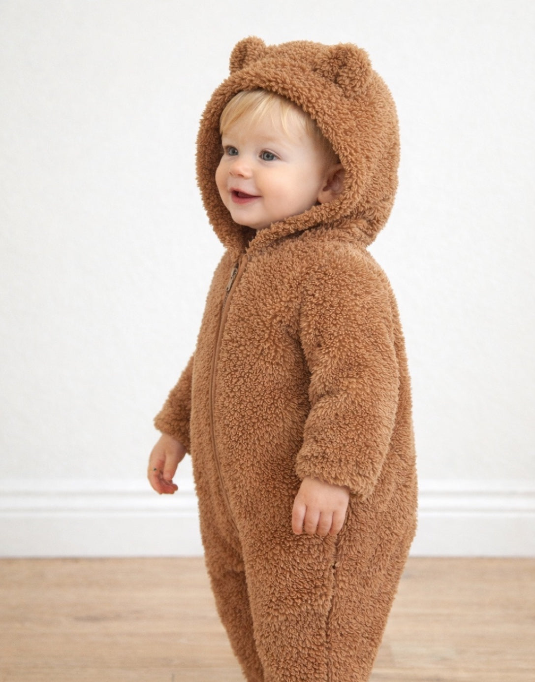 Side view of child wearing a brown fleece onesie with bear ears against a white wall.