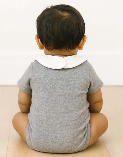 Back view of baby wearing a gray onesie with a white collar sitting on a light-colored surface.