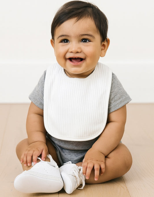 Baby wearing a white bib sitting on a white surface with a neutral background
