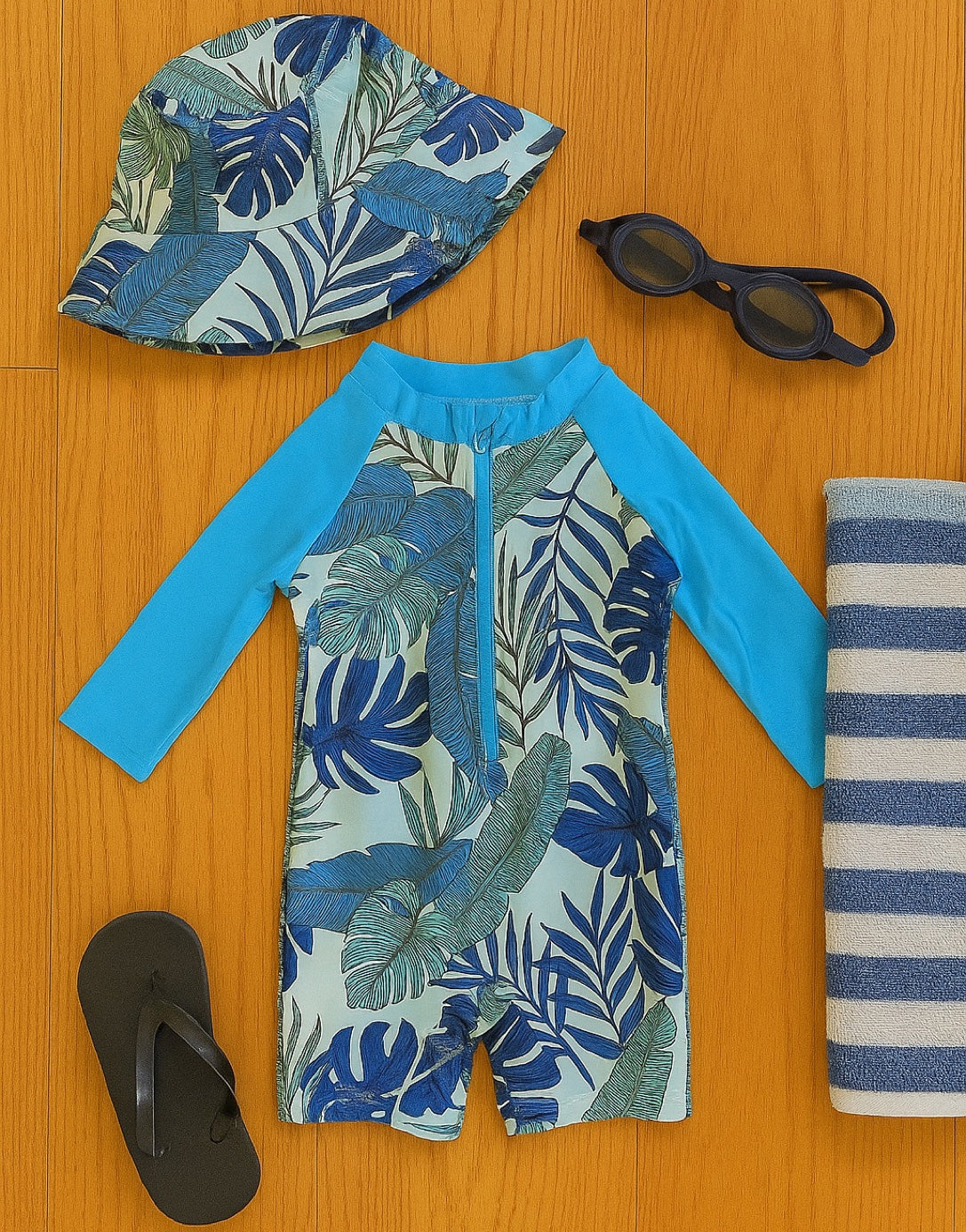 Blue and white leaf-patterned Baby Boy Zipper Front One Piece Swimsuit with a matching bucket hat, black sandal, swim goggles and striped beach towel.