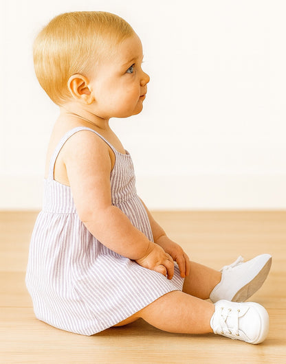 Side view of baby sitting on a wooden floor wearing a striped Baby Girl Bow Front Cami Dress and white shoes.
