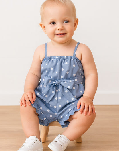 Baby wearing a blue polka dot Cami Ruffle Hem Romper with a bow on a wooden stool against a white background