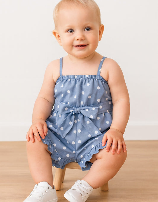 Baby wearing a blue polka dot Cami Ruffle Hem Romper with a bow on a wooden stool against a white background