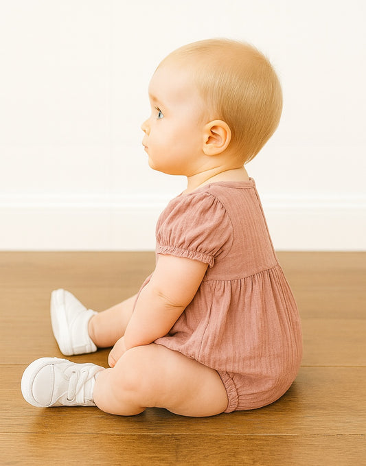 Side view of baby sitting on a wooden floor wearing a pink Puff Sleeve Onesie Romper and white shoes.