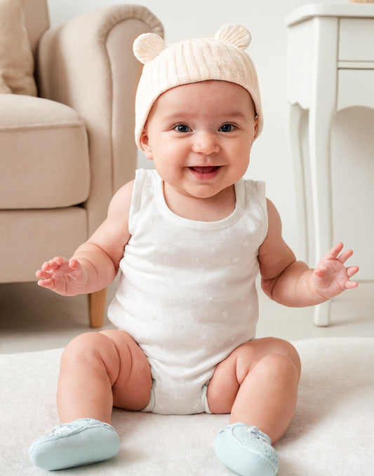 A baby wearing a white Hat with cute bear ears, sitting on a cozy white surface.