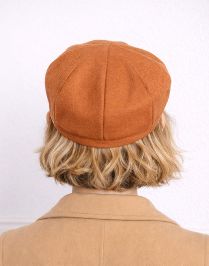 Back view of woman wearing a brown Baker Boy Cap and coat against a white background.