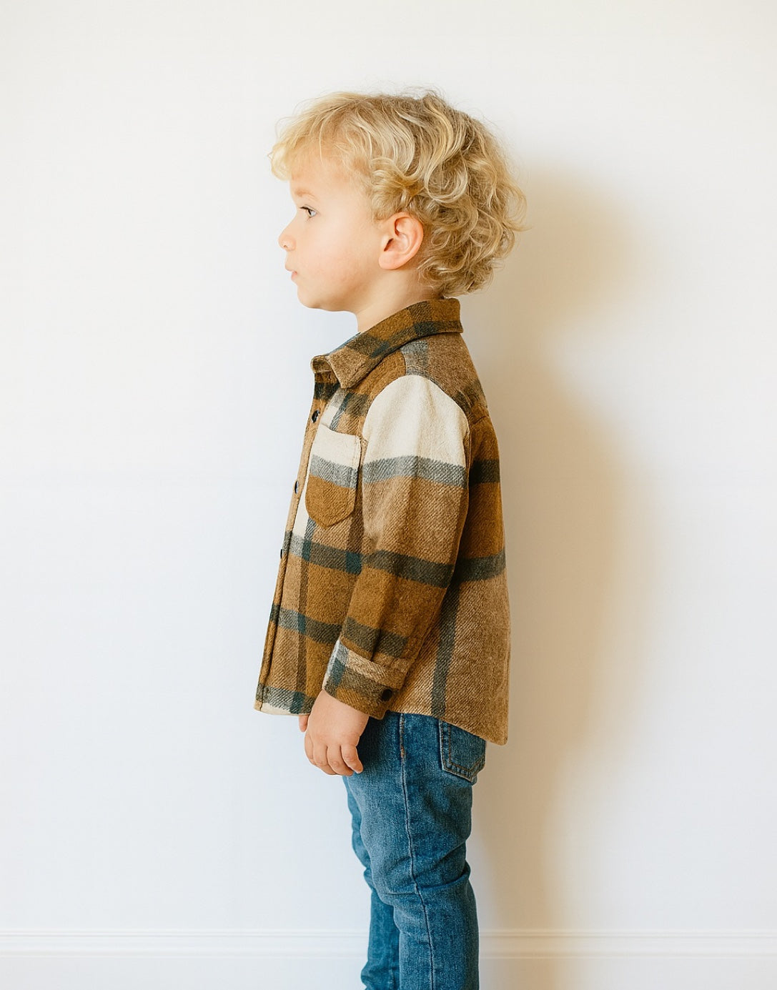 Side view of young boy wear brown and white plaid Shirt Jacket with denim jeans in front of plain white background.