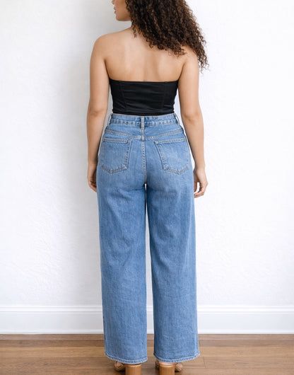 Back view of woman wearing a black strapless Bustier Crop Top and blue jeans against a white wall.