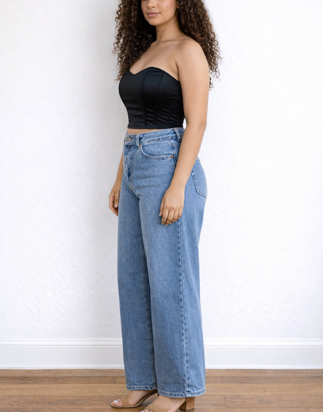 Side view of woman wearing a black strapless Bustier Crop Top and blue jeans against a white wall.