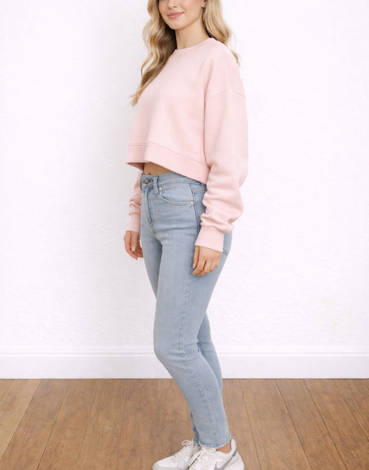 Side view of woman wearing a pink Crop Sweatshirt and light blue jeans against a white wall.