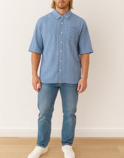 Man wearing a blue checkered Drop Shoulder Loose Fit Button Down Shirt and jeans standing against a white wall.