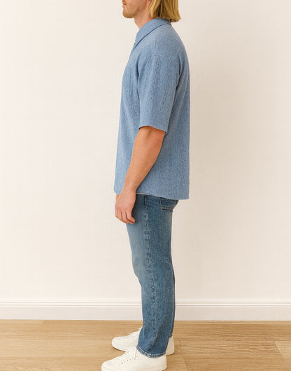 Side view of person wearing a blue denim Drop Shoulder Loose Fit Button Down Shirt and jeans standing against a white wall.