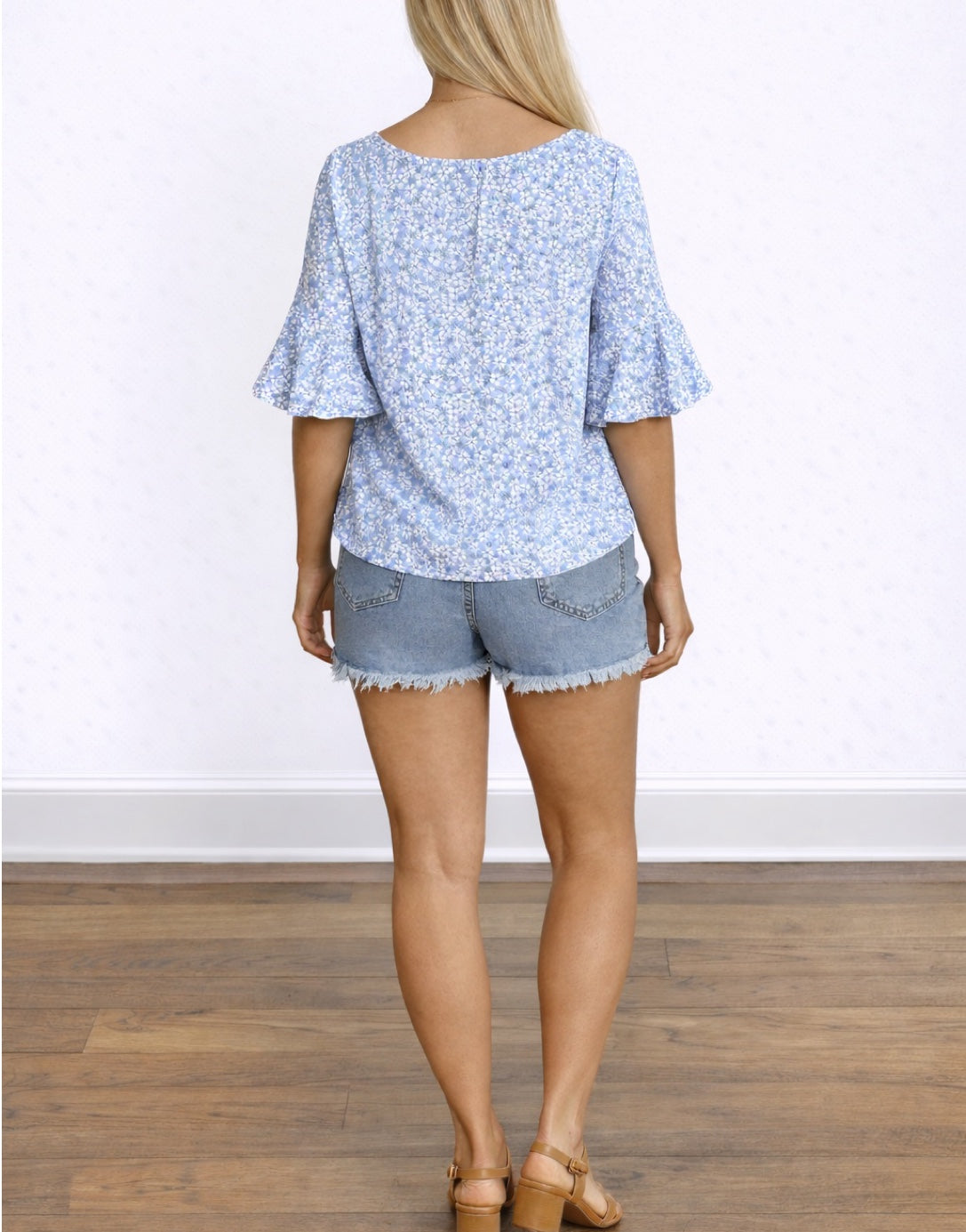 Back view of woman wearing a blue floral Flounce Sleeve Blouse and denim shorts on a white background.