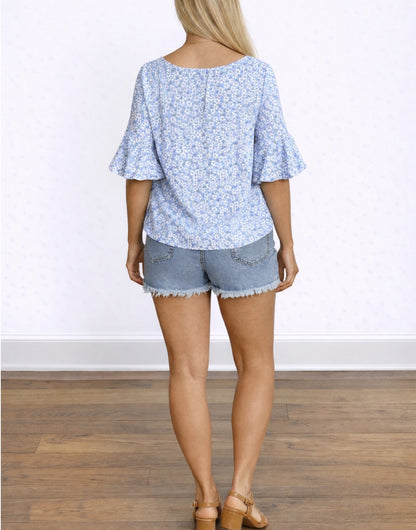 Back view of woman wearing a blue floral Flounce Sleeve Blouse and denim shorts on a white background.
