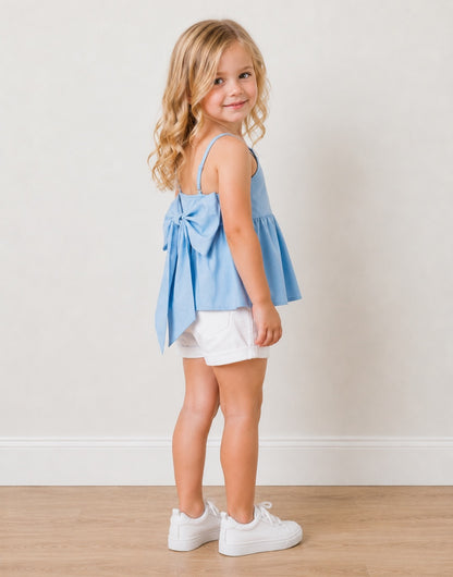Side view of young girl wearing a light blue Bow Back Babydoll Cami Top and white shorts standing against a plain background.