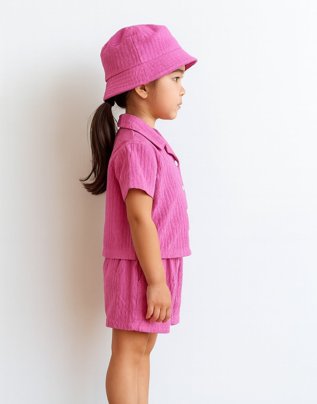 Side view of child wearing a pink Button Front Shirt and Shorts Set with a matching hat on a white background.