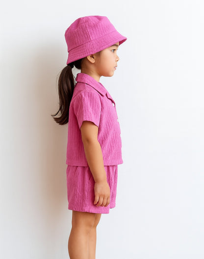 Side view of child wearing a pink Button Front Shirt and Shorts Set with a matching hat on a white background.