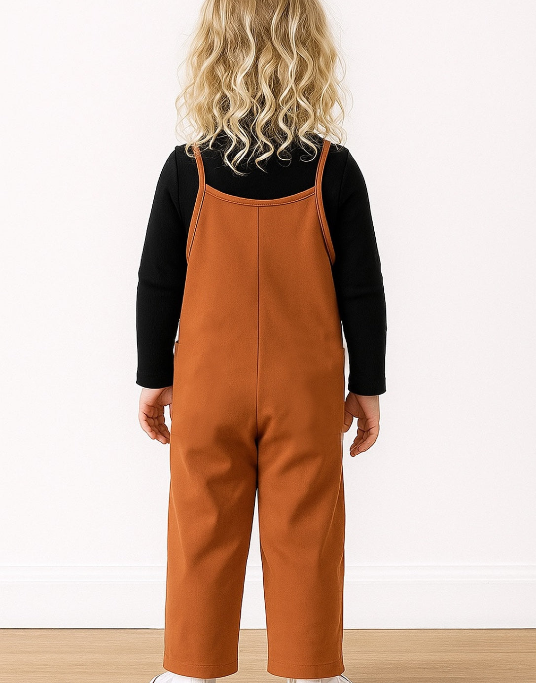 Back view of child wearing a brown overalls and black shirt against a white background.