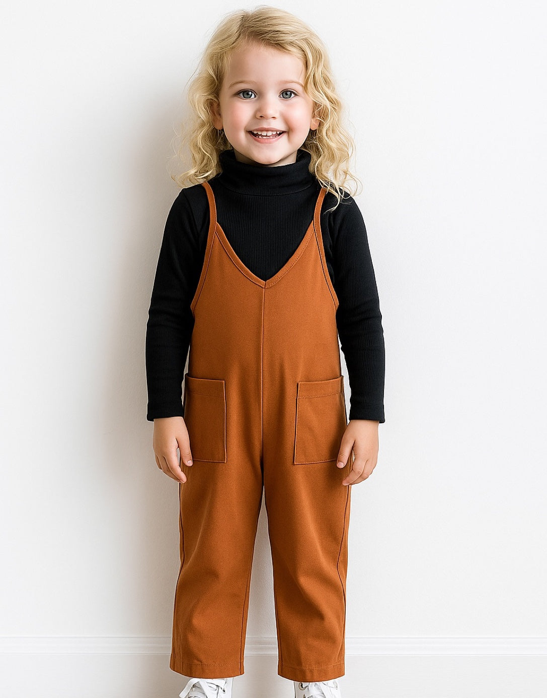 Child wearing brown Cami Knit Jumpsuit standing on steps with a white wall and greenery in the background