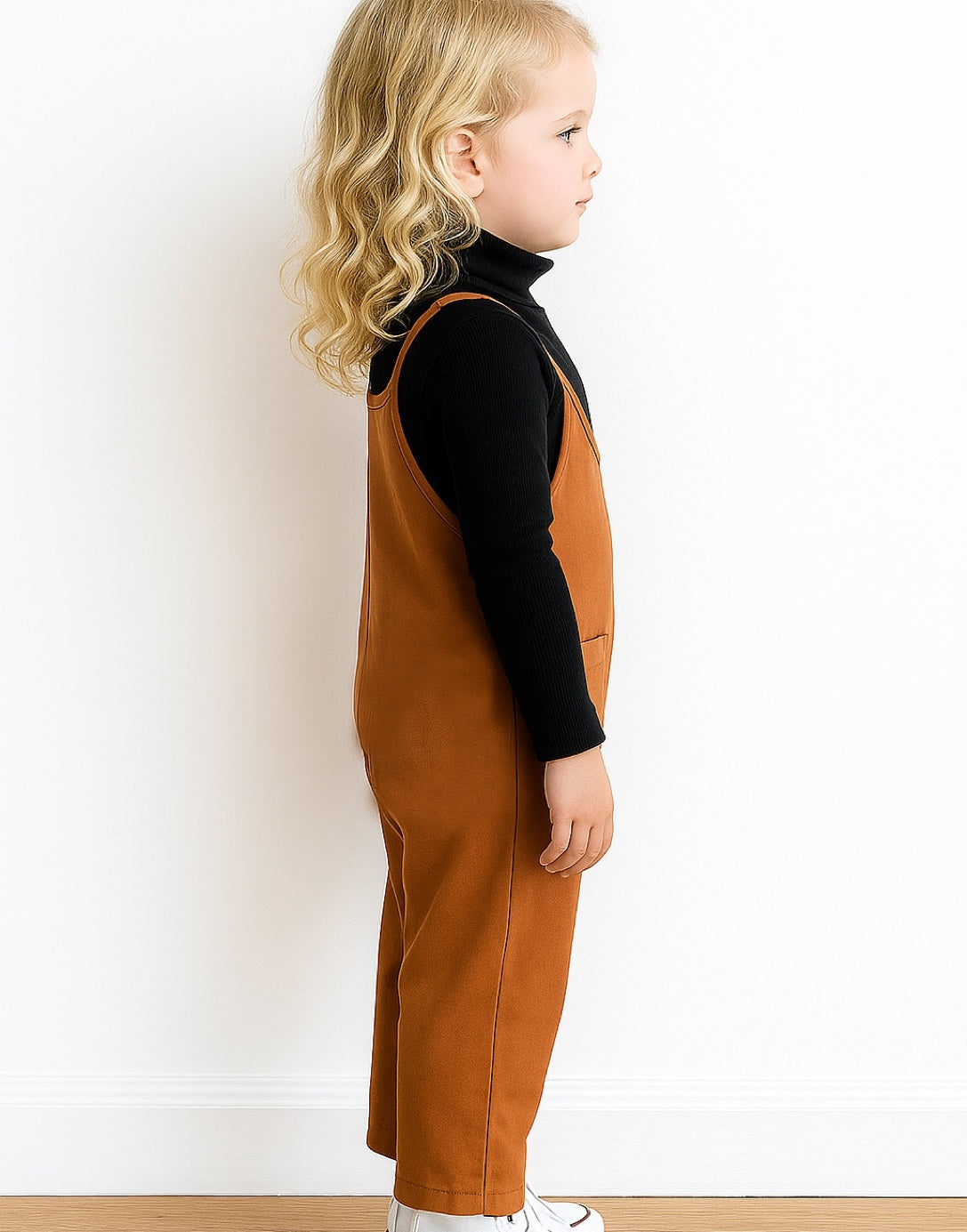 Side view of child wearing a brown overalls and black shirt against a white background.