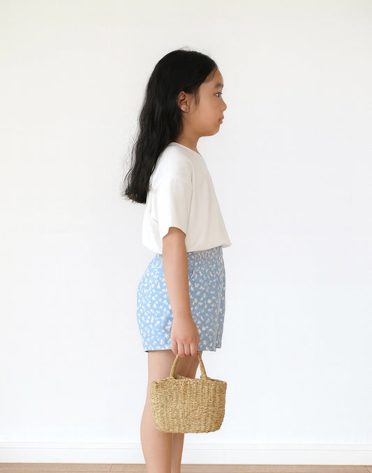 Side view of young girl wearing a white top and light blue patterned Elastic Shirred Waist Shorts, holding a woven bag against a plain background.