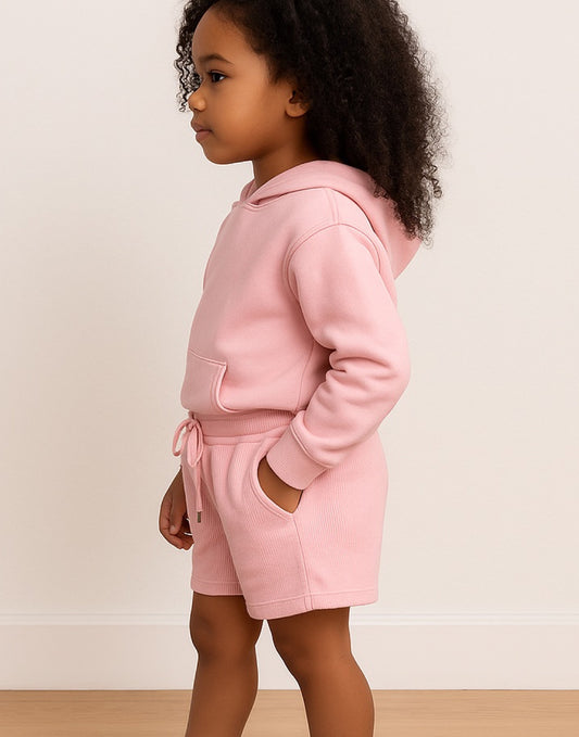 Side view of young girl wearing a white top and pink Gym Shorts against a beige wall.