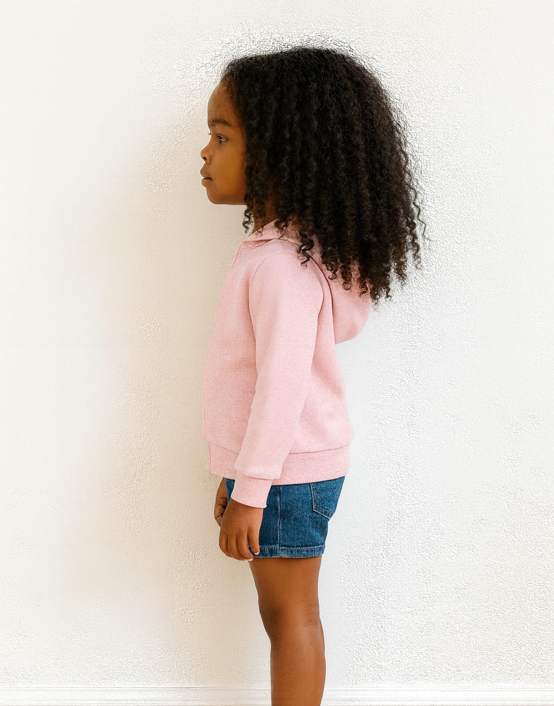 Side view of young girl wearing pink Zip Up Hoodie Jacket and denim jeans in front of white wall background and wood floor.