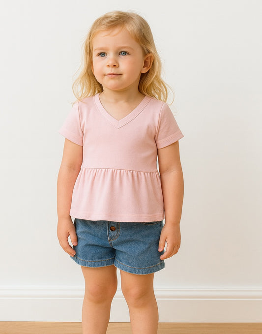 Young girl wearing a pink knit peplum top and blue shorts against a white wall.