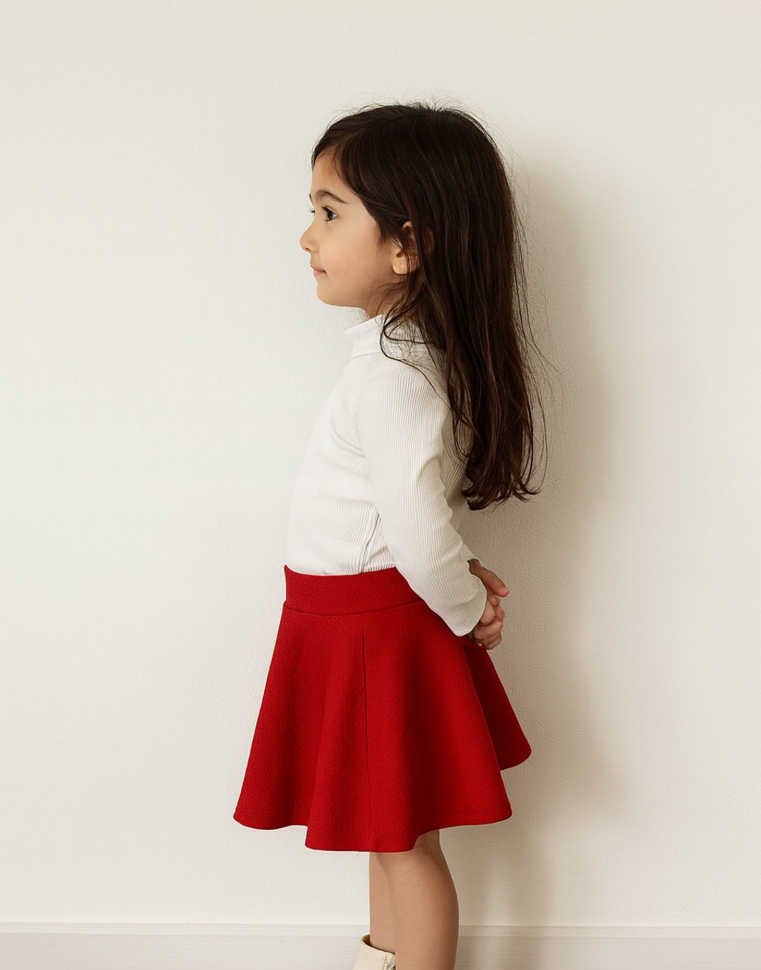Side view of young girl wearing a white sweater and red Mini Circle Skirt against a white background.