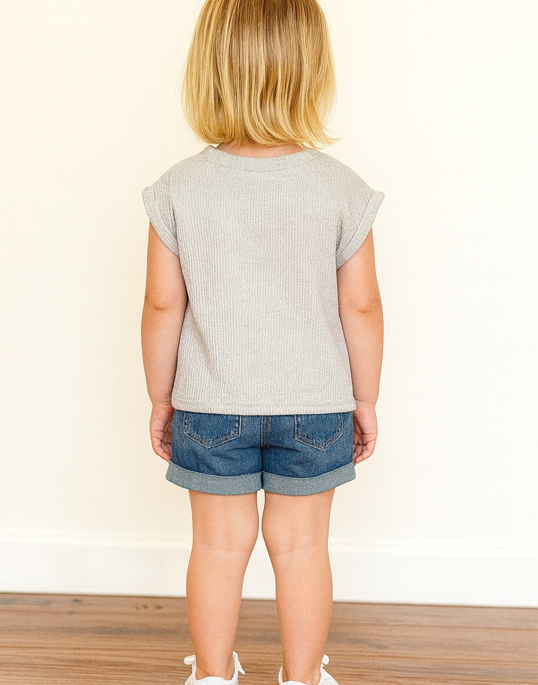 Back view of young girl wearing a grey Oversized Tee Shirt with denim shorts and white shoes in front of a white wall with wooden floor.