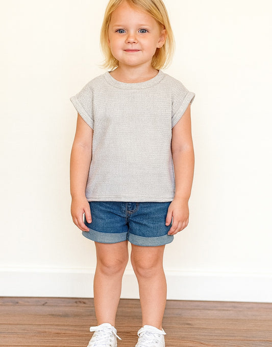 Young girl wearing a grey Oversized Tee with denim shorts and white shoes in front of a white wall with wooden floor