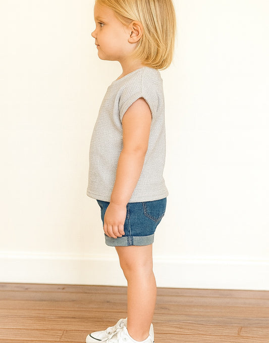 Side view of young girl wearing a grey Oversized T Shirt with denim shorts and white shoes in front of a white wall with wooden floor.