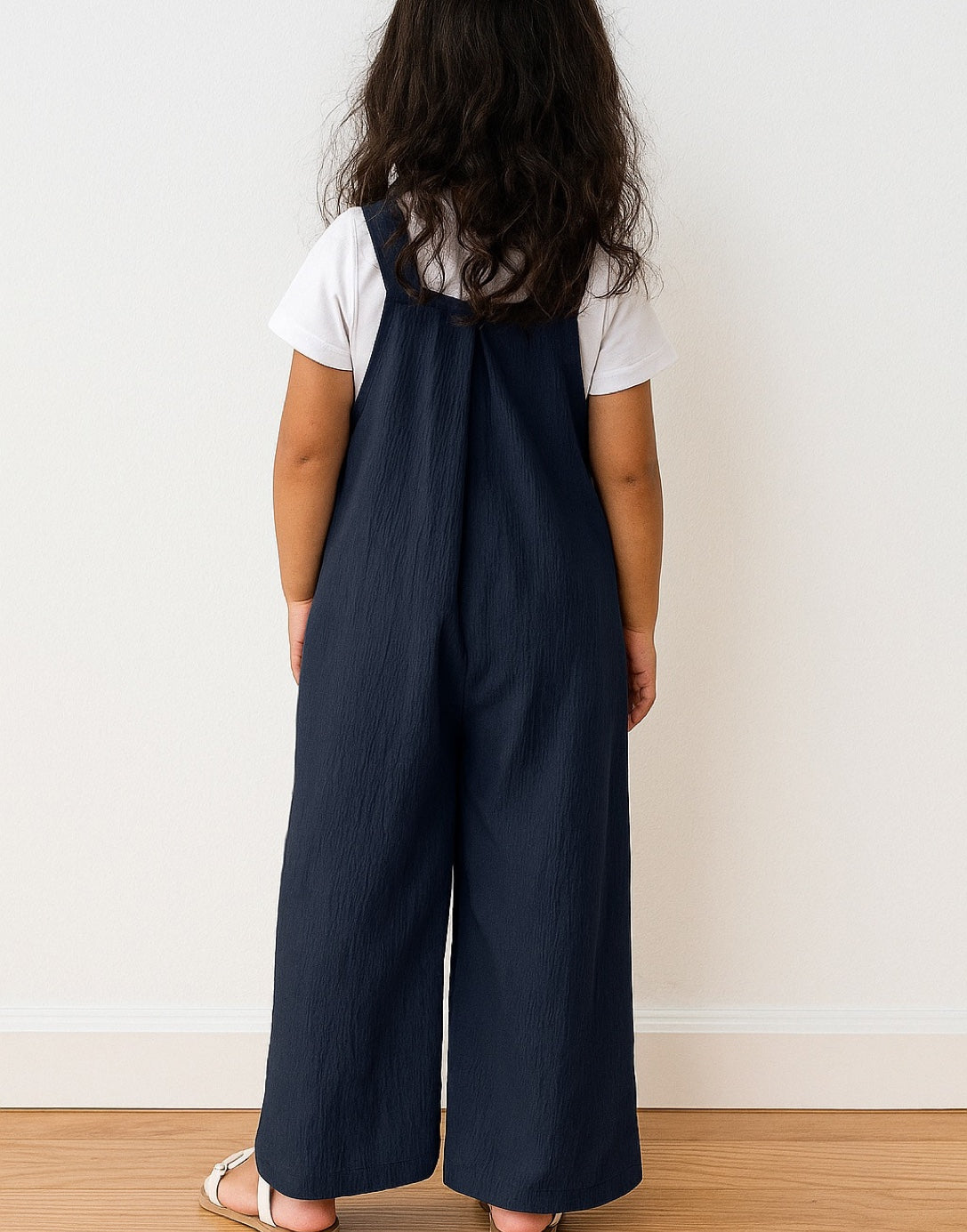 Back view of young girl wearing navy blue Pinafore Jumpsuit and a white shirt against a white wall.