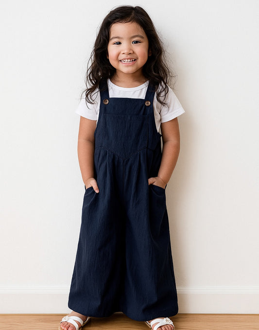 Young girl wearing navy blue Pinafore Jumpsuit and a white shirt against a white wall.