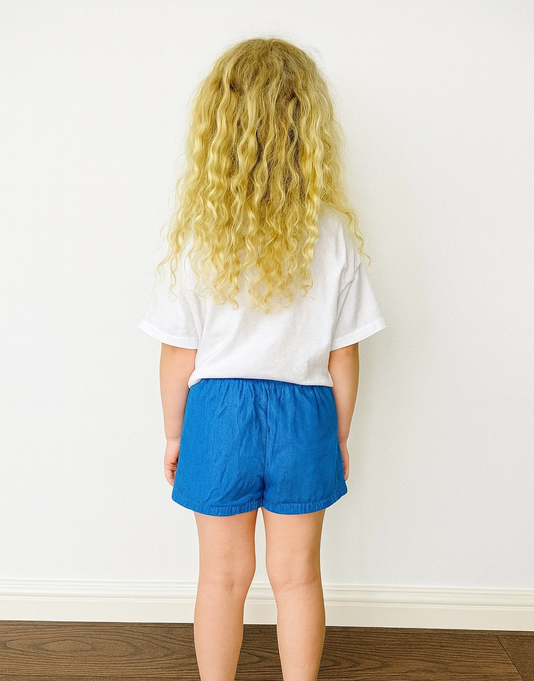 Back view of young girl wearing a white t-shirt and blue Ruffle Hem Bow Skort against a plain background.