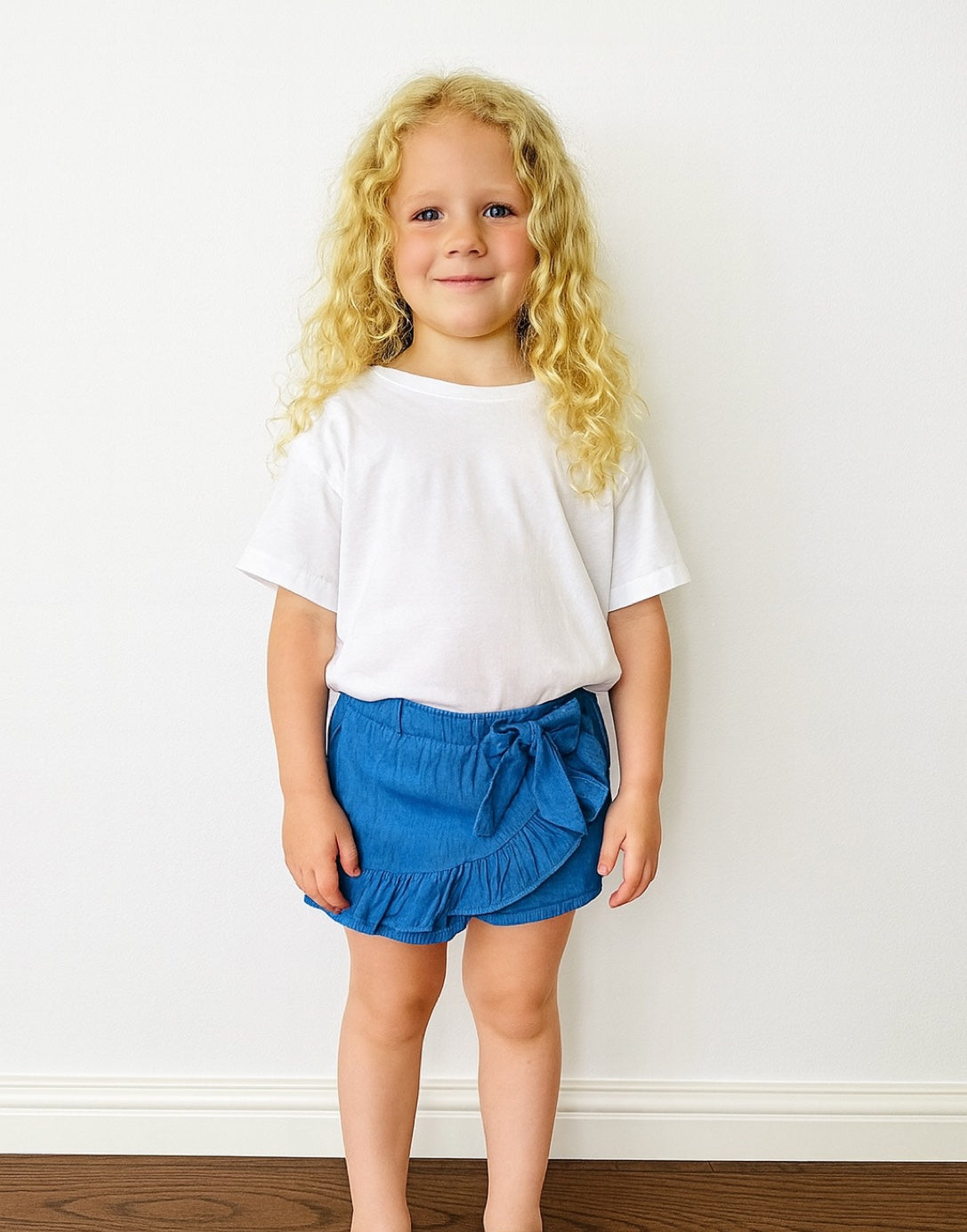 Young girl wearing a white t-shirt and blue Ruffle Hem Bow Skort against a plain background