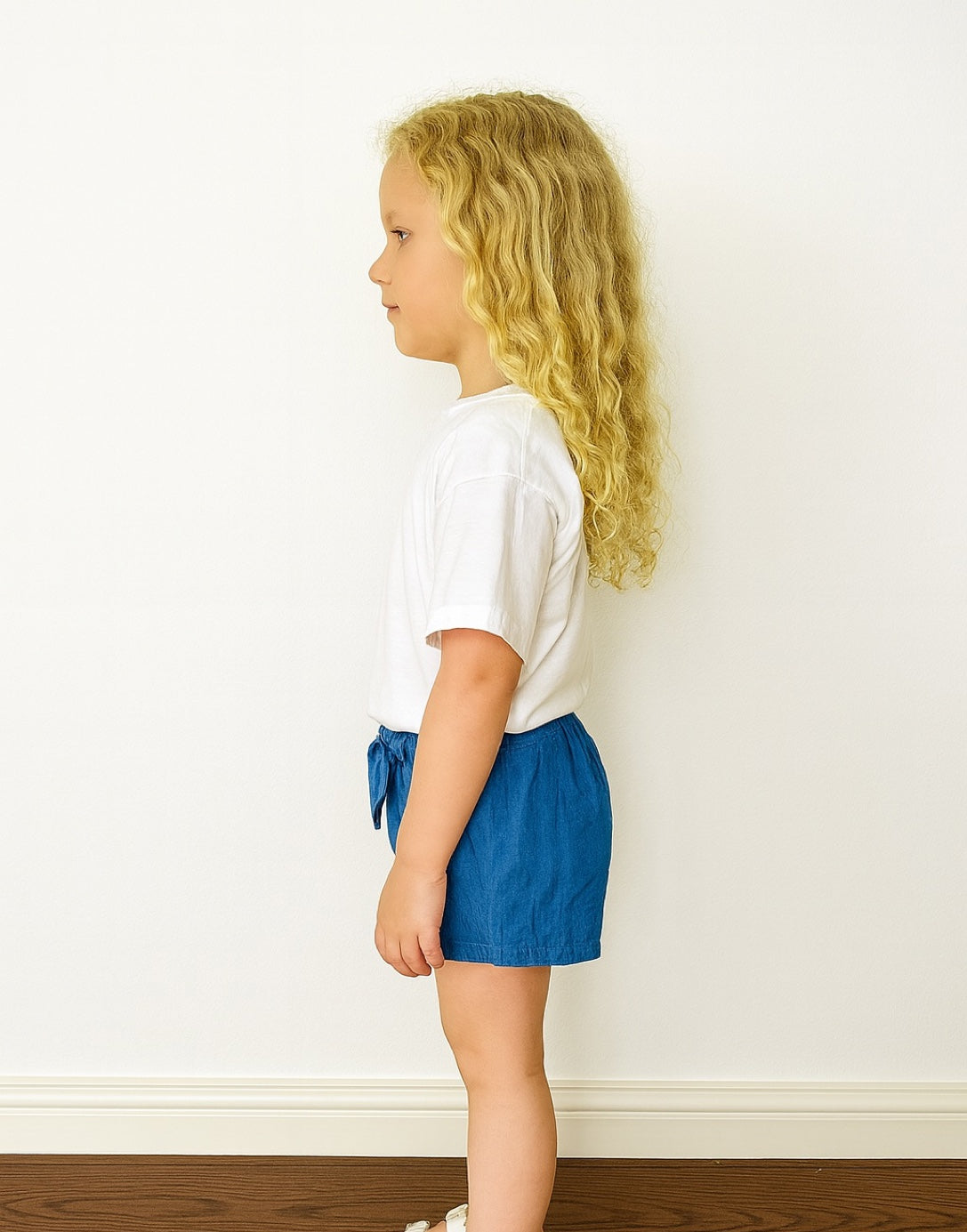 Side view of young girl wearing a white t-shirt and blue Ruffle Hem Bow Skort against a plain background.
