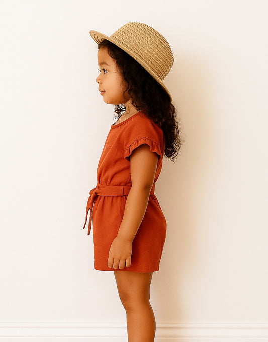 Side view of child wearing a rust-colored Ruffle Sleeve Belted Romper and straw hat against a white background.