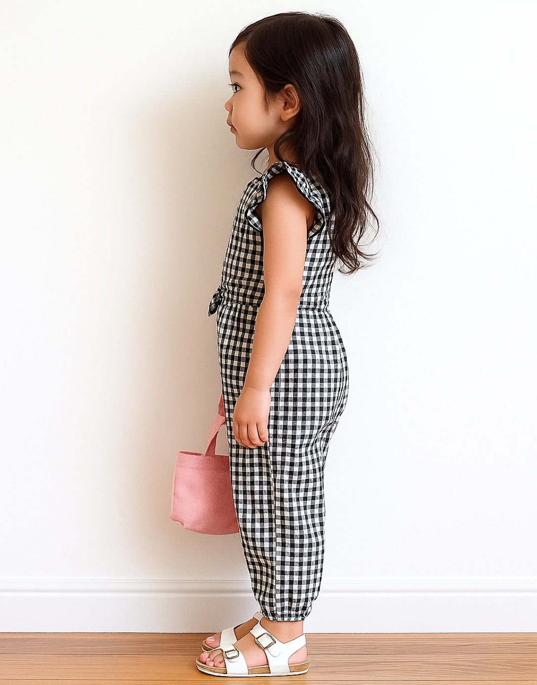 Side view of young girl wearing a black and white checkered Ruffle Sleeve Jumpsuit holding a pink bag against a plain background.