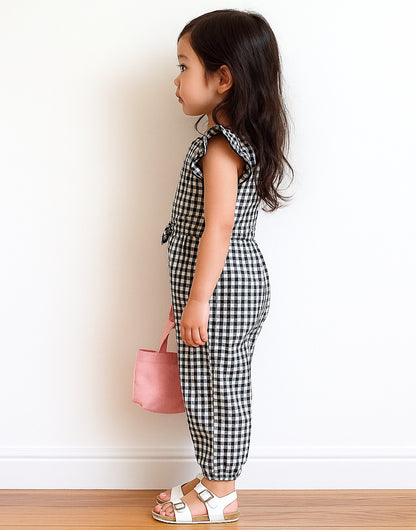 Side view of young girl wearing a black and white checkered Ruffle Sleeve Jumpsuit holding a pink bag against a plain background.