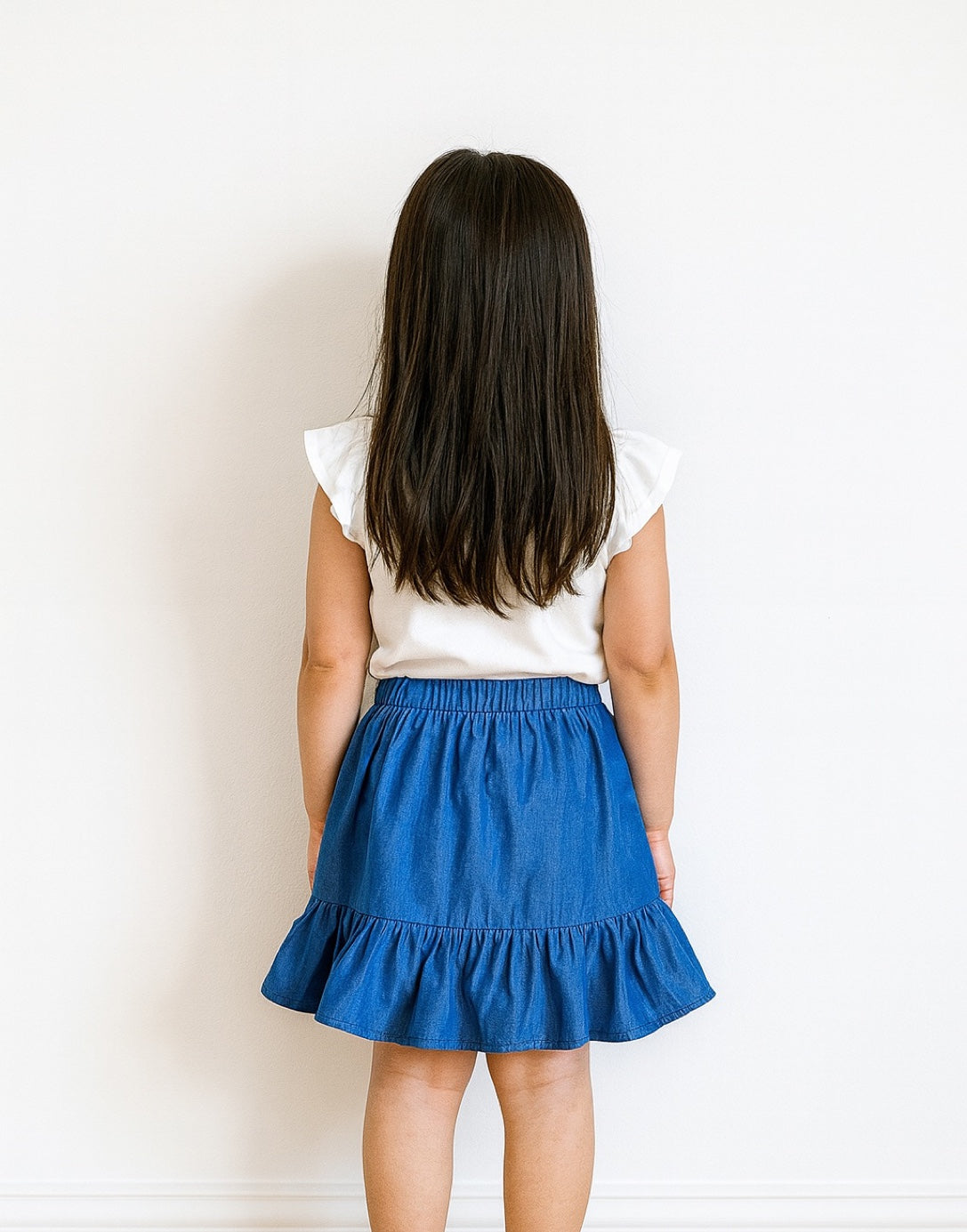 Back view of young girl wearing a white top and blue Ruffle Wrap Mini Skirt against a plain background.
